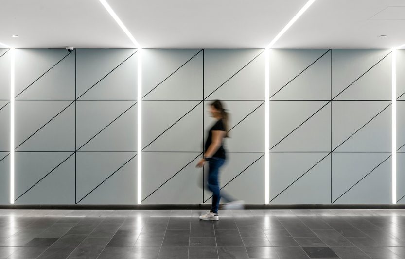 Persona sfocata cammina davanti a parete moderna con luci a LED diagonali in un ambiente farmaceutico high-tech.