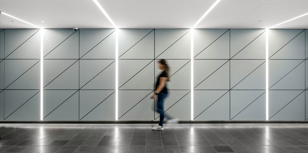 Persona sfocata cammina davanti a parete moderna con luci a LED diagonali in un ambiente farmaceutico high-tech.