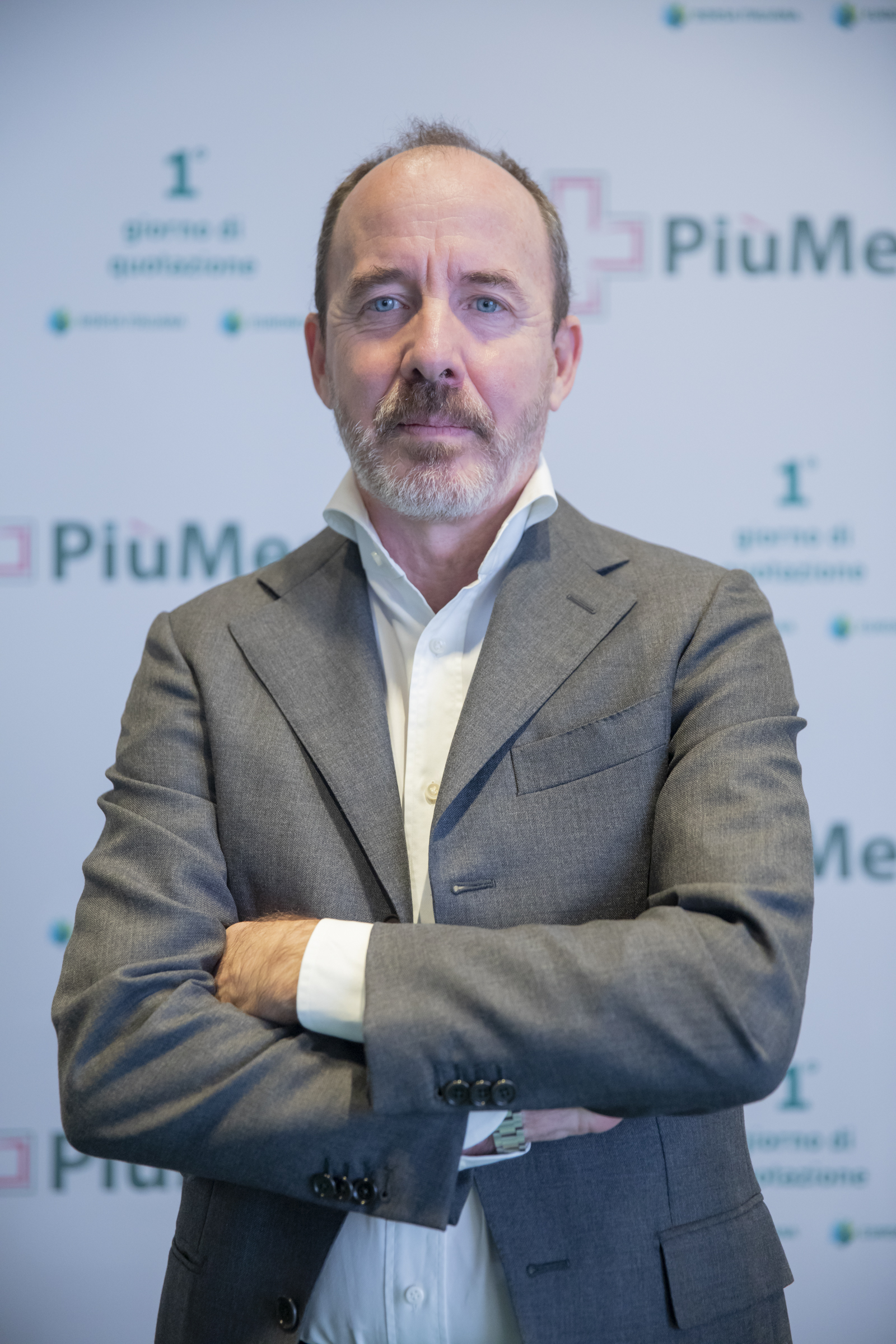 Uomo in giacca grigia davanti a un logo di PiuMe, settore farmaceutico. Sfondo con testo sfocato e dettagli aziendali.