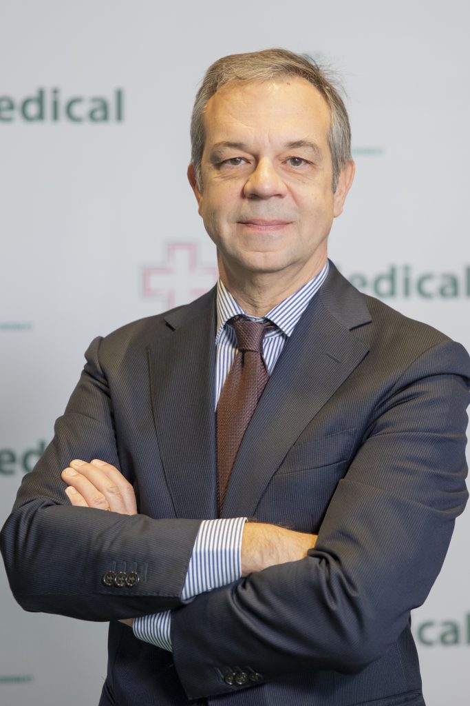 Uomo maturo in abito formale con braccia incrociate, sfondo con logo medico sfocato.