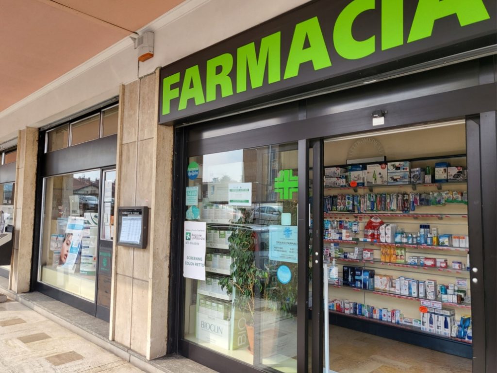 Ingresso di una farmacia con vetrina espositiva di prodotti medici e insegna luminosa verde.