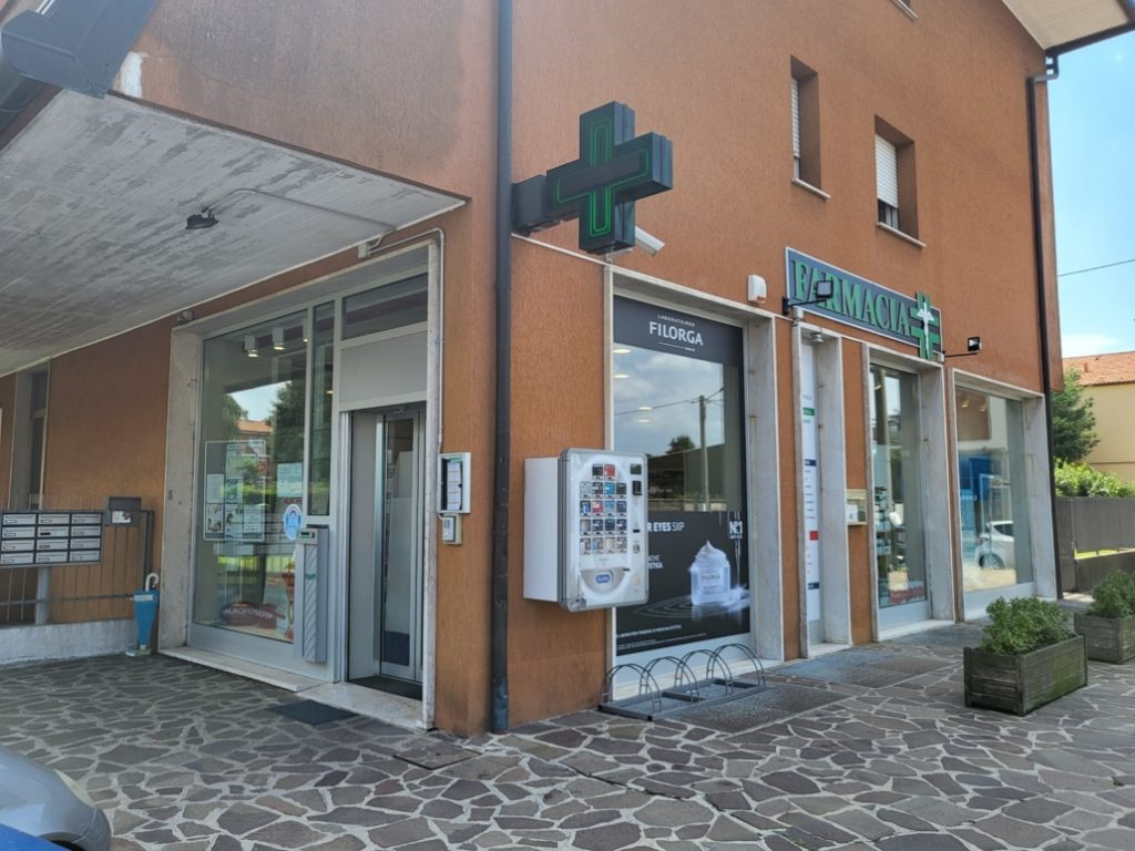 Farmacia moderna con insegna a croce verde luminosa e distributore automatico esterno su facciata al sole.