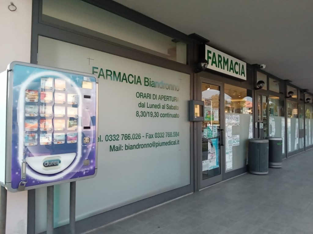 Ingresso di una farmacia con distributore automatico di preservativi, orari di apertura e contatti visibili sulla vetrina.