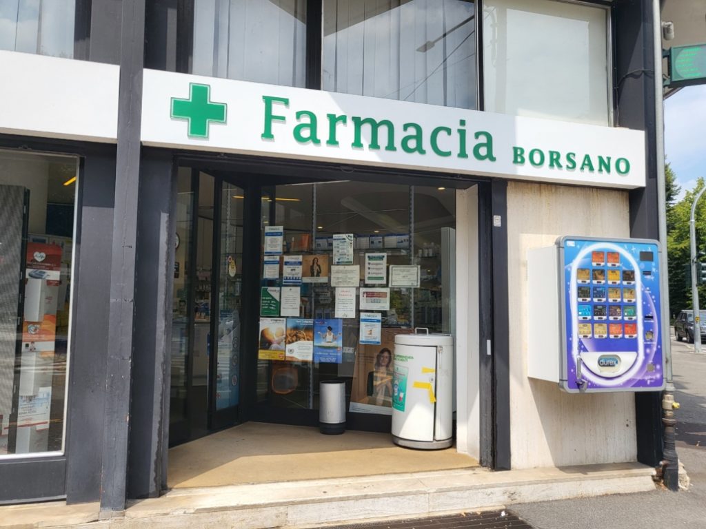 Ingresso di una farmacia con distributore automatico Durex all'esterno. Cartelli e prodotti visibili all'interno.