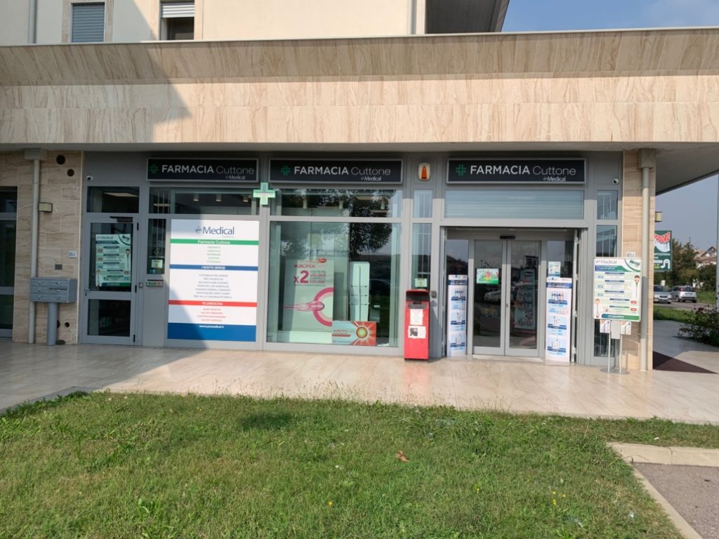 Facciata di una farmacia moderna con insegne visibili, vetrine trasparenti e manifesti promozionali.