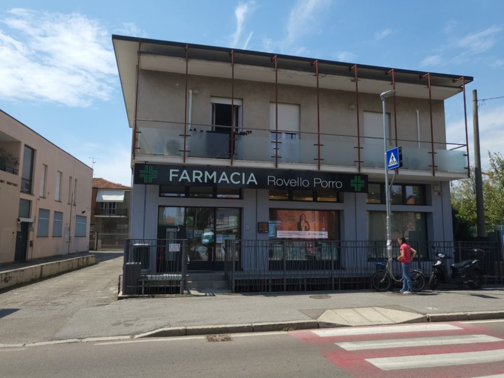 Facciata di una farmacia moderna con insegne verdi, situata in una zona urbana, con persone e biciclette fuori.
