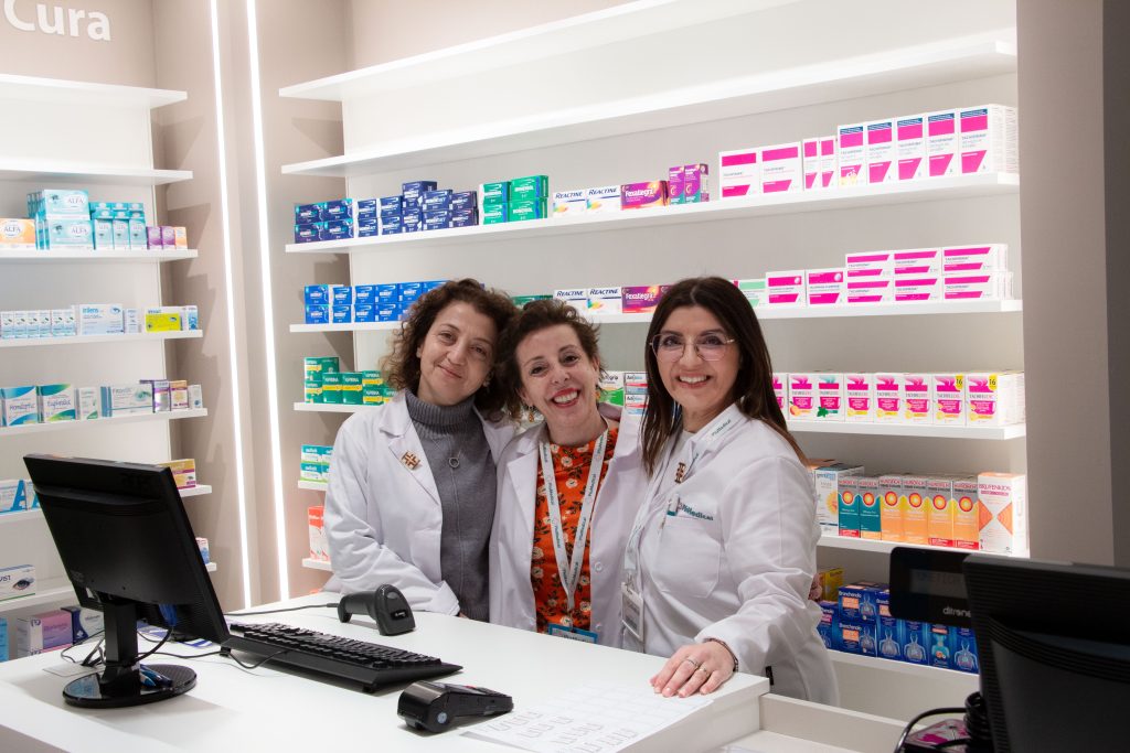 Pharmacy staff smiling behind the counter with a variety of medication displayed on shelves.