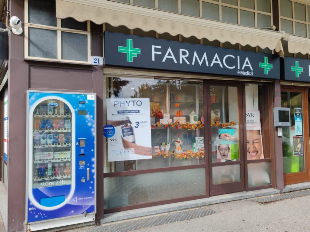 Facciata di una farmacia con insegna visibile e distributore automatico di prodotti sanitari accanto.