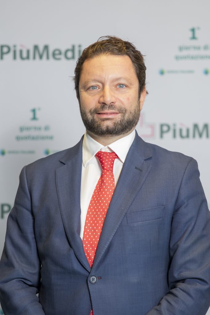 Uomo in giacca e cravatta rossa, sfondo con loghi PiuMedico, evento del settore farmaceutico.